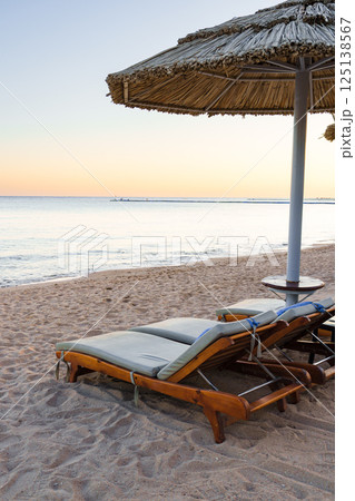Relaxing beachside loungers under a straw umbrella at sunset by the calm ocean waves Relaxing beachside loungers under a straw umbrella at sunset by the calm ocean waves 125138567