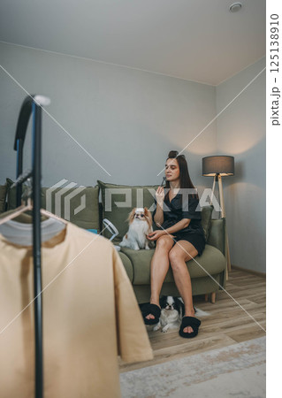 woman in a black bathrobe, relaxing on a green sofa care of her face, massage of face while savoring the company of her two adorable dogs, clothes rack 125138910