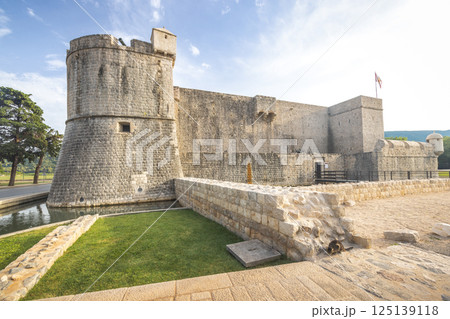 Ston, historic town in Peljesac peninsula in Croatia, Europe. Ancient stone castle with a round tower, surrounded by a moat and grassy area under a bright blue sky with scattered clouds. 125139118
