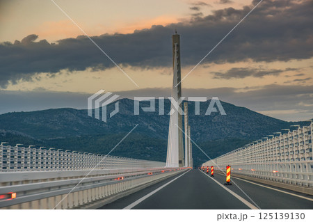 The Peljesac Bridge to Peljesac peninsula in Croatia, Europe. Driving across a modern bridge at dusk, supported by tall pillars against a backdrop of cloudy skies and distant mountains. The Peljesac Bridge to Peljesac peninsula in Croatia, Europe. Driving across a modern bridge at dusk, supported by tall pillars against a backdrop of cloudy skies and distant mountains. 125139130