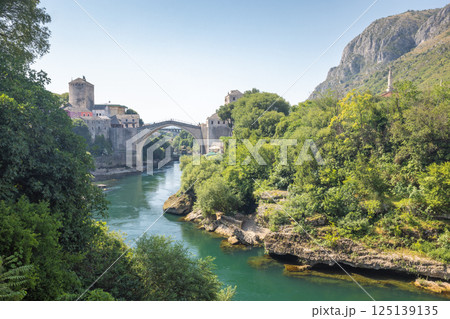 The Stari Most bridge, old bridge on Neretva river in Mostar town, Bosnia and Herzegovina, Europe. Scenic view of a stone bridge and river surrounded by lush greenery, old buildings, and mountains. 125139135