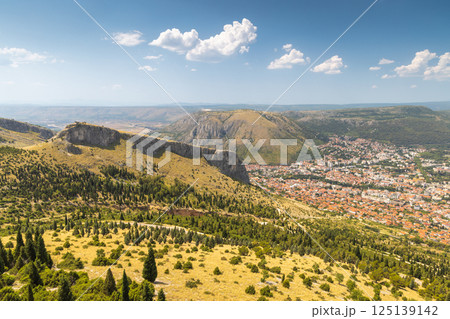 View of Mostar from the Fortica hill, Bosnia and Herzegovina, Europe. Sweeping aerial view captures a charming town nestled within a mountainous landscape under a sunny, partly cloudy sky. View of Mostar from the Fortica hill, Bosnia and Herzegovina, Europe. Sweeping aerial view captures a charming town nestled within a mountainous landscape under a sunny, partly cloudy sky. 125139142