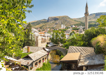 Mostar, historic town in Bosnia and Herzegovina, Europe. Picturesque old town scene with a stone bridge, tiled roofs, and a distant minaret beneath a blue sky in a European setting. 125139146