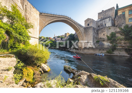 The Stari Most bridge, old bridge on Neretva river in Mostar town, Bosnia and Herzegovina, Europe. Picturesque stone bridge stretching over a vibrant river with a boat gliding underneath. The Stari Most bridge, old bridge on Neretva river in Mostar town, Bosnia and Herzegovina, Europe. Picturesque stone bridge stretching over a vibrant river with a boat gliding underneath. 125139166