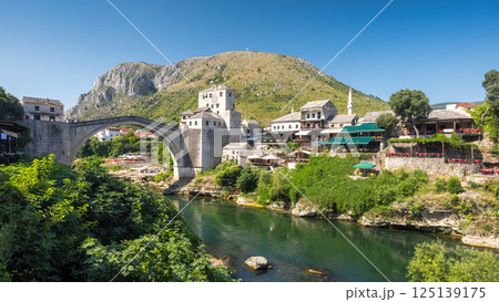 The Stari Most bridge, old bridge on Neretva river in Mostar town, Bosnia and Herzegovina, Europe. Iconic bridge spanning tranquil river with historic buildings, lush greenery, and mountain backdrop. 125139175