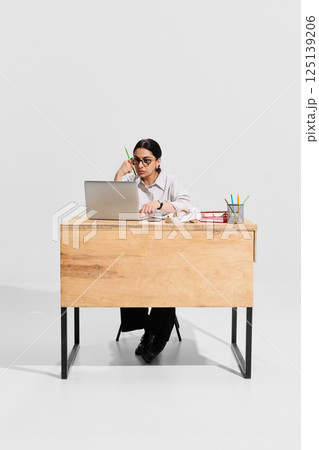Focused woman, teacher in white blouse sitting at desk with stationary and book, working on laptop and preparing for the lessons on white background. 125139206