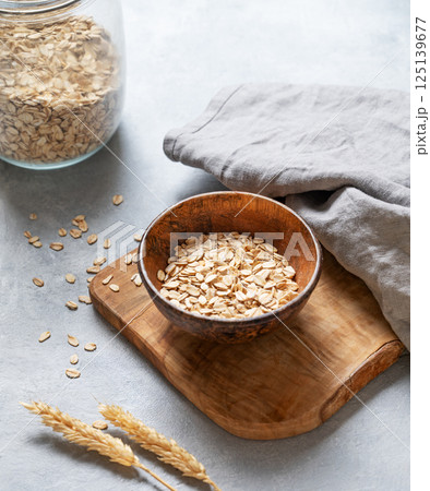 Oat flakes in bowl on wooden board with napkin, wheat and glass jar on grey textured background. 125139677