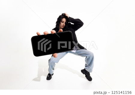 Stylish woman in black blazer and jeans strikes expressive pose, stretching hand with smartphone toward camera against white studio background. 125141156