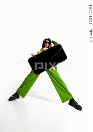 Woman in green with hand extended holding smartphone directly toward viewer against white studio background. 125141161