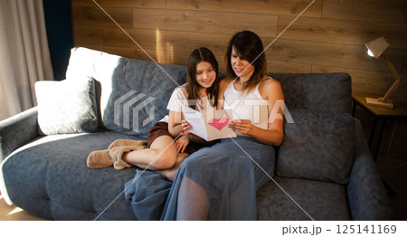 Mom and daughter celebrating mother's day handmade reading greeting card sitting on sofa in home interior. Mom and daughter celebrating mother's day handmade reading greeting card sitting on sofa in home interior. 125141169