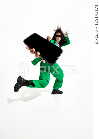 Stylish woman in bright green suit and sunglasses lunges forward with smartphone with blank screen against white background. Stylish woman in bright green suit and sunglasses lunges forward with smartphone with blank screen against white background. 125141173