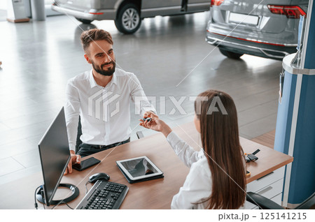 Sitting by the table and taking the keys. Man with woman in white clothes are in the car dealership together 125141215