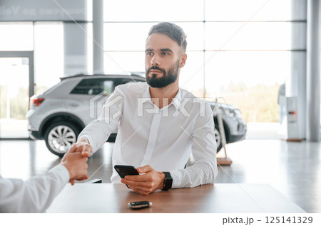 Sitting by the table. Young man in white clothes is in the car dealership 125141329