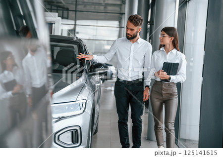 Touching the automobile surface. Man with woman in white clothes are in the car dealership together Touching the automobile surface. Man with woman in white clothes are in the car dealership together 125141615