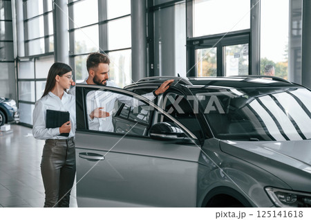 Touching the automobile surface. Man with woman in white clothes are in the car dealership together Touching the automobile surface. Man with woman in white clothes are in the car dealership together 125141618