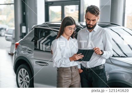 Black tablet is in the hands. Reading information. Man with woman in white clothes are in the car dealership together Black tablet is in the hands. Reading information. Man with woman in white clothes are in the car dealership together 125141620