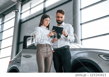 Black tablet is in the hands. Reading information. Man with woman in white clothes are in the car dealership together Black tablet is in the hands. Reading information. Man with woman in white clothes are in the car dealership together 125141621