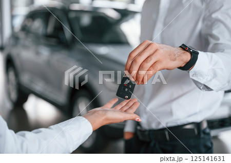 Keys of new automobile. Close up view. Man with woman in white clothes are in the car dealership together Keys of new automobile. Close up view. Man with woman in white clothes are in the car dealership together 125141631