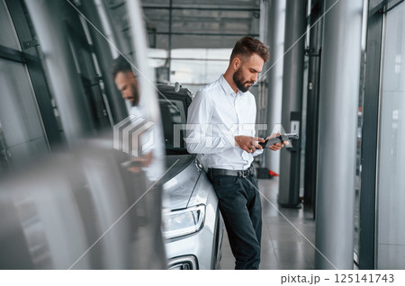 Black graphic tablet in hands. Young man in white clothes is in the car dealership 125141743