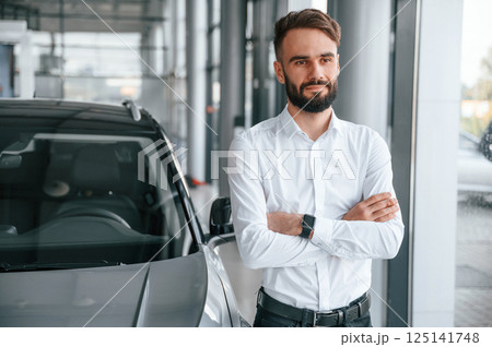 Standing with arms crossed. Young man in white clothes is in the car dealership 125141748