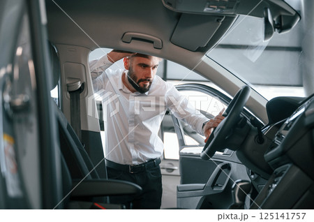 Examining the salon of the automobile. Young man in white clothes is in the car dealership Examining the salon of the automobile. Young man in white clothes is in the car dealership 125141757