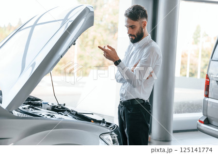 Looks under the hood of automobile. Young man in white clothes is in the car dealership Looks under the hood of automobile. Young man in white clothes is in the car dealership 125141774