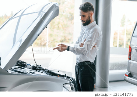 Looks under the hood of automobile. Young man in white clothes is in the car dealership Looks under the hood of automobile. Young man in white clothes is in the car dealership 125141775