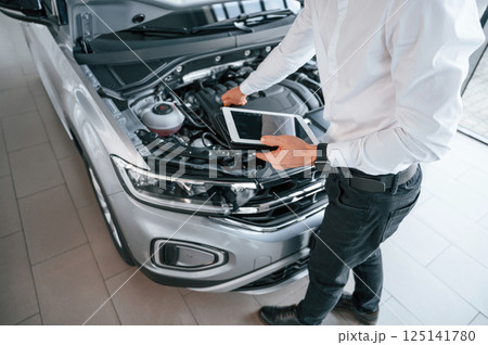 Looking under the hood and holding tablet. Young man in white clothes is in the car dealership Looking under the hood and holding tablet. Young man in white clothes is in the car dealership 125141780