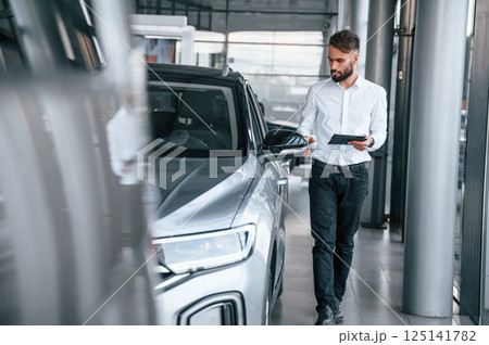 Standing and holding black graphic tablet in hand. Young man in white clothes is in the car dealership Standing and holding black graphic tablet in hand. Young man in white clothes is in the car dealership 125141782