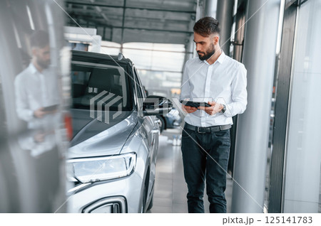 Standing and holding black graphic tablet in hand. Young man in white clothes is in the car dealership Standing and holding black graphic tablet in hand. Young man in white clothes is in the car dealership 125141783