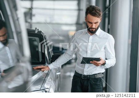 Standing with table in hands. Young man in white clothes is in the car dealership Standing with table in hands. Young man in white clothes is in the car dealership 125142211