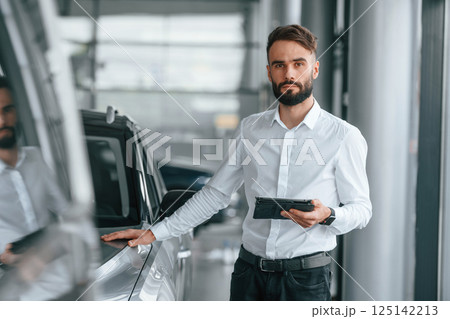 Touching the surface of automobile. Young man in white clothes is in the car dealership 125142213