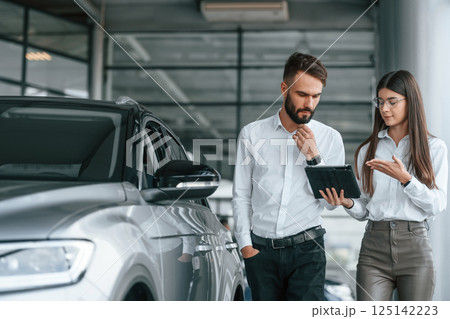Using digital tablet. Man with woman in white clothes are in the car dealership together 125142223