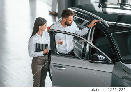 Opening the door. Man with woman in white clothes are in the car dealership together 125142234