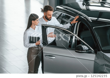 Opening the door. Man with woman in white clothes are in the car dealership together 125142235