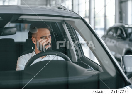 Front view. Sitting inside of automobile. Young man in white clothes is in the car dealership 125142279
