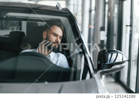 Front view. Sitting inside of automobile. Young man in white clothes is in the car dealership Front view. Sitting inside of automobile. Young man in white clothes is in the car dealership 125142280