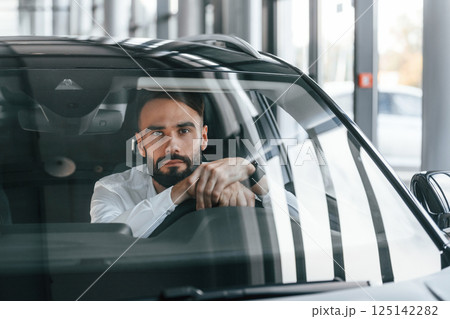 Front view. Sitting inside of automobile. Young man in white clothes is in the car dealership 125142282