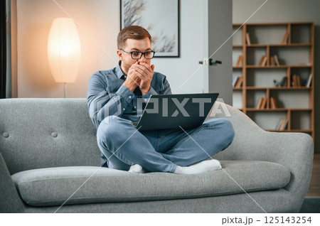 Feeling shocked. Male freelance worker is using laptop at home. Cozy apartment Feeling shocked. Male freelance worker is using laptop at home. Cozy apartment 125143254