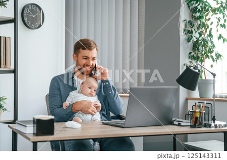 Sitting by the table with laptop. Father with toddler is indoors in the office Sitting by the table with laptop. Father with toddler is indoors in the office 125143311