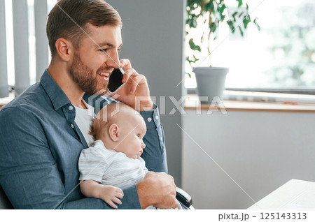 Talking by phone. Father with toddler is indoors in the office 125143313