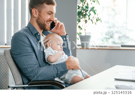 Talking by phone. Father with toddler is indoors in the office 125143316