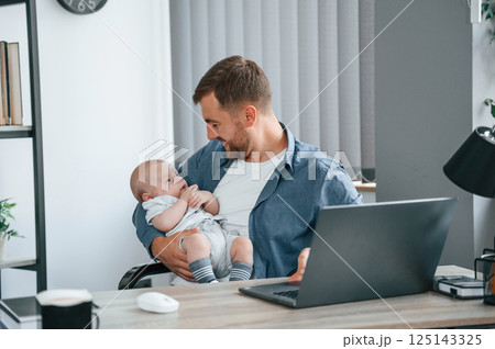 Working by using laptop. Holding baby. Father with toddler is indoors in the office 125143325