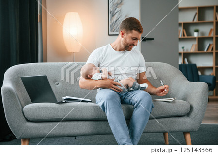 Sitting on the sofa and using pen. Father with toddler is at home, taking care of his son Sitting on the sofa and using pen. Father with toddler is at home, taking care of his son 125143366