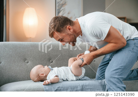 Baby is laying down on the sofa. Father with toddler is at home, taking care of his son Baby is laying down on the sofa. Father with toddler is at home, taking care of his son 125143390