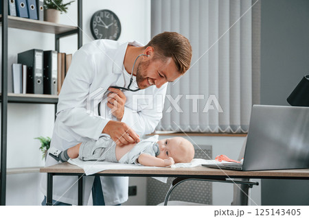 Using stethoscope. Doctor in white coat is taking care of little toddler indoors 125143405