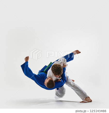 High-speed shoulder throw in progress, athlete in white executes flawless lift, blue gi fighter suspended mid-air, representing victory against white background. 125143466
