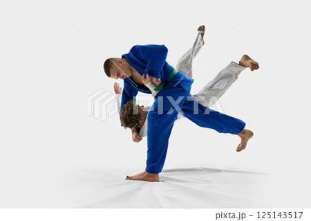Judoka in white executes forward throw, lifting opponent over shoulder in full motion, reflecting strength, precision against white studio background. Judoka in white executes forward throw, lifting opponent over shoulder in full motion, reflecting strength, precision against white studio background. 125143517