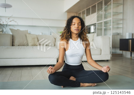Yoga mindfulness meditation. Young healthy African girl practicing yoga at home. Woman sitting in lotus pose on yoga mat meditating relaxing indoor. Girl doing breathing practice. Yoga at home 125143546