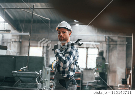 Standing and holding wrench. Factory male worker in uniform is indoors Standing and holding wrench. Factory male worker in uniform is indoors 125143711